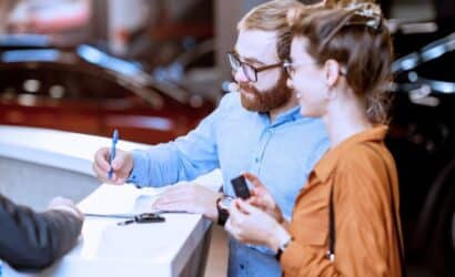 A man and woman at a dealership counter, signing documents and receiving keys for a vehicle purchase.
